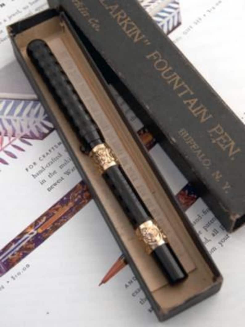 Puede incluir: Una pluma estilogr&aacute;fica negra vintage con detalles dorados, presentada en su caja original. La pluma tiene un cuerpo texturizado y detalles ornamentados. La caja es oscura con el texto "LARKIN" FOUNTAIN PEN" y "BUFFALO, N.Y."