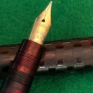 Puede incluir: Primer plano de una pluma estilogr&aacute;fica vintage con plum&iacute;n dorado. El plum&iacute;n est&aacute; marcado "Warranted 14K S.A.". El cuerpo de la pluma es de color marr&oacute;n oscuro con un dise&ntilde;o texturizado. El fondo es una superficie verde.