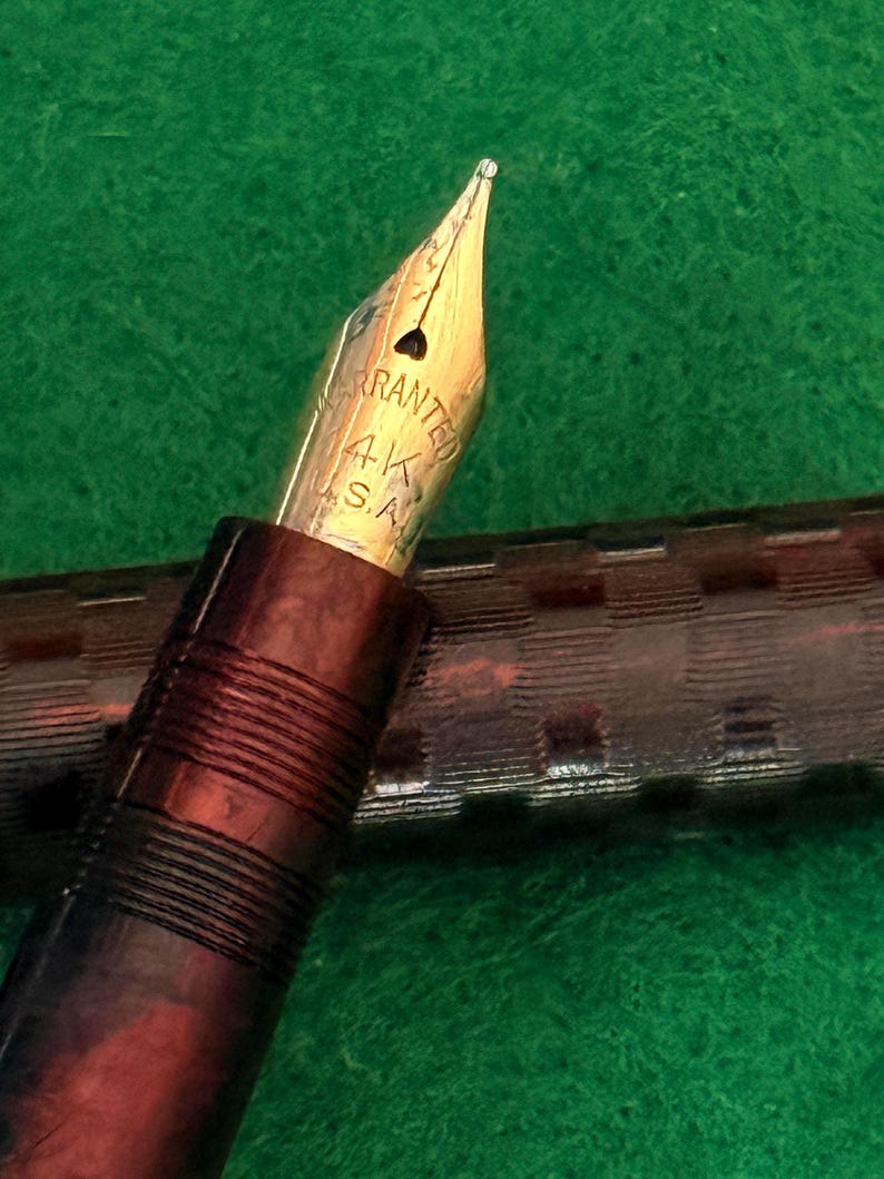 Puede incluir: Primer plano de una pluma estilogr&aacute;fica vintage con plum&iacute;n dorado. El plum&iacute;n est&aacute; marcado "Warranted 14K S.A.". El cuerpo de la pluma es de color marr&oacute;n oscuro con un dise&ntilde;o texturizado. El fondo es una superficie verde.