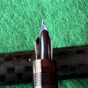 Puede incluir: Primer plano del plum&iacute;n de una pluma estilogr&aacute;fica. El plum&iacute;n es plateado y marr&oacute;n, con una punta fina. El cuerpo de la pluma es marr&oacute;n oscuro con un dise&ntilde;o acanalado. El fondo es una superficie verde texturizada.