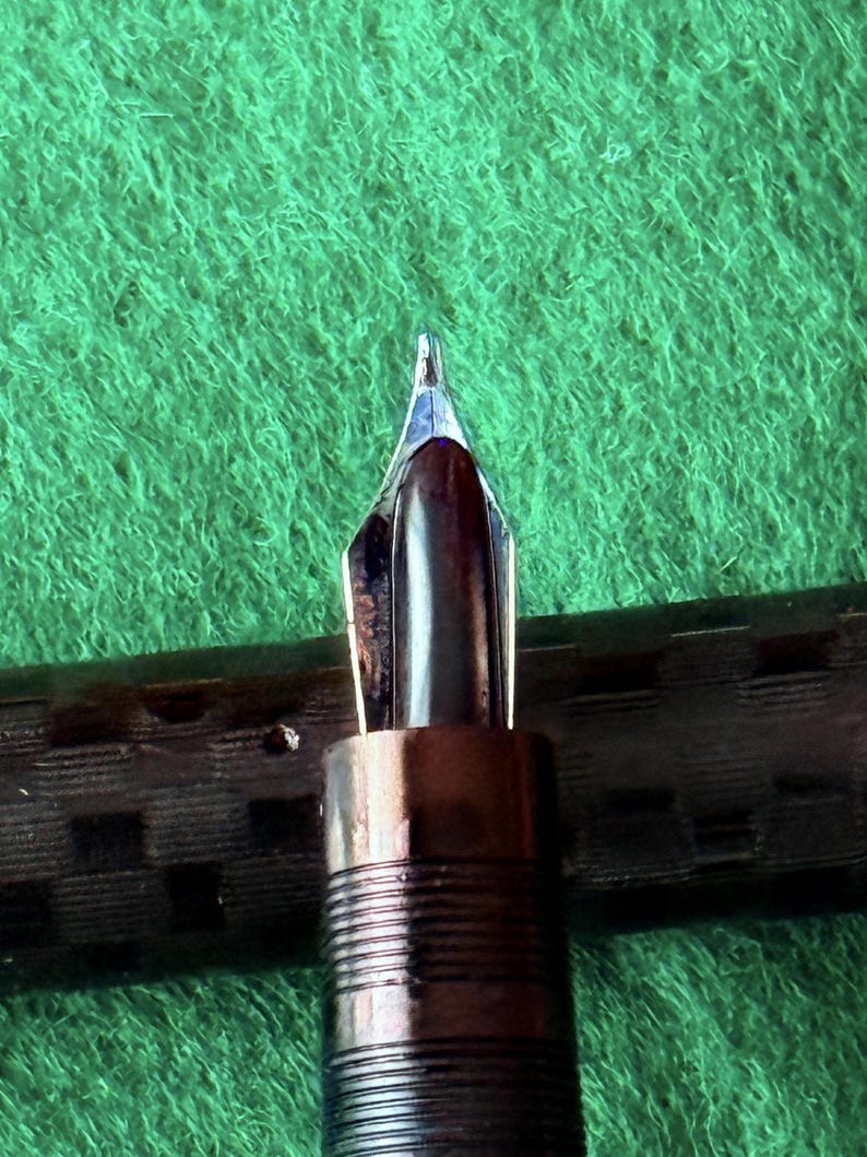 Puede incluir: Primer plano del plum&iacute;n de una pluma estilogr&aacute;fica. El plum&iacute;n es plateado y marr&oacute;n, con una punta fina. El cuerpo de la pluma es marr&oacute;n oscuro con un dise&ntilde;o acanalado. El fondo es una superficie verde texturizada.