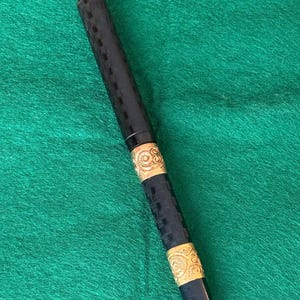 Puede incluir: Un bol&iacute;grafo negro con bandas decorativas doradas. El cuerpo principal del bol&iacute;grafo tiene un patr&oacute;n texturizado y a cuadros. Las bandas doradas presentan un dise&ntilde;o ornamental y arremolinado. El bol&iacute;grafo est&aacute; sobre un fondo verde.