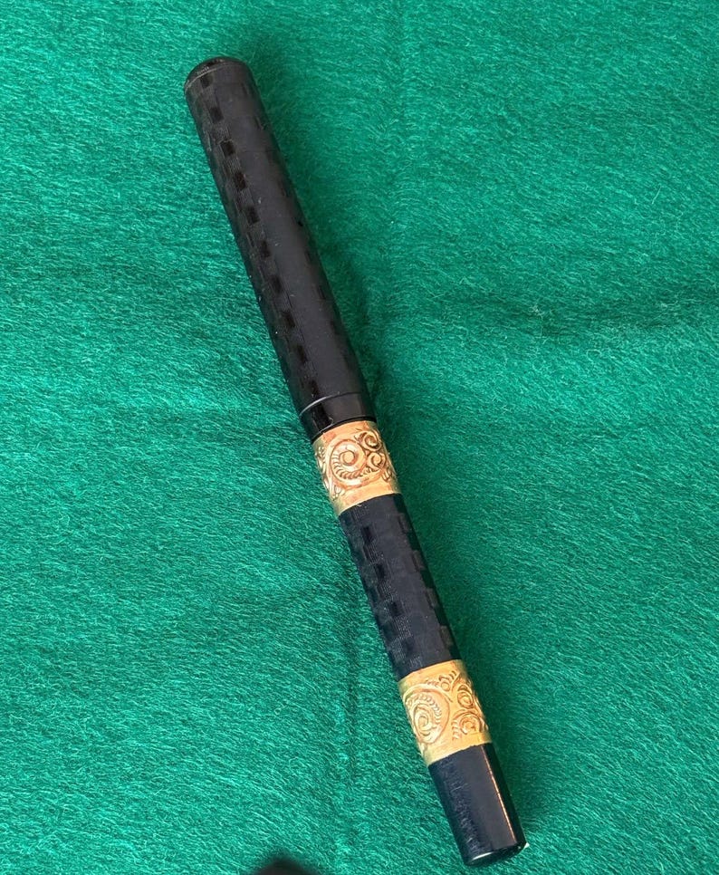 Puede incluir: Un bol&iacute;grafo negro con bandas decorativas doradas. El cuerpo principal del bol&iacute;grafo tiene un patr&oacute;n texturizado y a cuadros. Las bandas doradas presentan un dise&ntilde;o ornamental y arremolinado. El bol&iacute;grafo est&aacute; sobre un fondo verde.