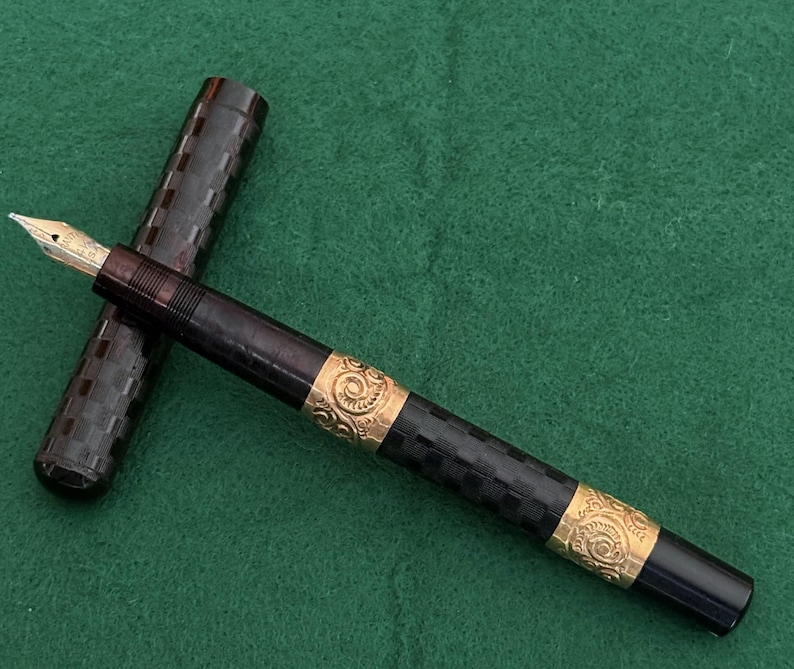 Puede incluir: Una pluma estilogr&aacute;fica vintage con plum&iacute;n de oro y bandas doradas ornamentadas. El cuerpo de la pluma es negro con un patr&oacute;n de cuadros, y la tapa est&aacute; separada. La pluma descansa sobre una superficie verde.