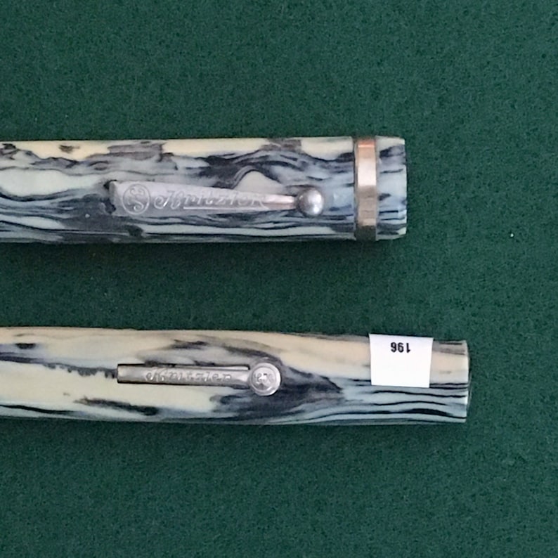 May include: Two black and white marbled pens with silver accents and the word "Schreiber" engraved on the side. The pen on the right has a gold band around the cap.