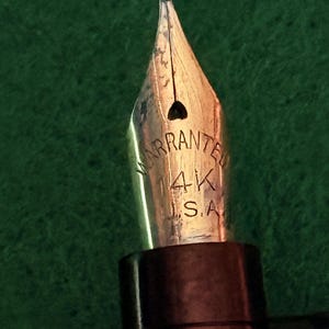 Puede incluir: Primer plano de un plum&iacute;n de pluma estilogr&aacute;fica dorado. El plum&iacute;n est&aacute; grabado con la palabra "WARRANTED" y las marcas "4K S.A.". La pluma tiene un cuerpo marr&oacute;n oscuro.