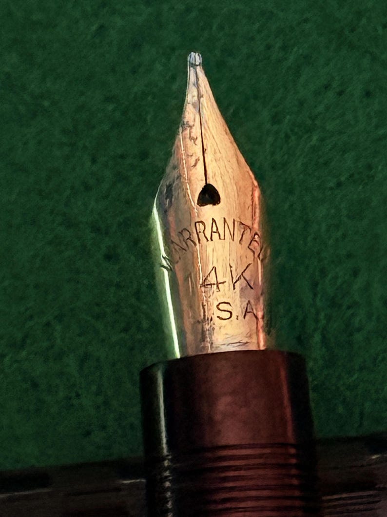 Puede incluir: Primer plano de un plum&iacute;n de pluma estilogr&aacute;fica dorado. El plum&iacute;n est&aacute; grabado con la palabra "WARRANTED" y las marcas "4K S.A.". La pluma tiene un cuerpo marr&oacute;n oscuro.