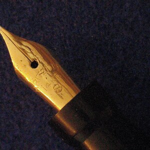 May include: A gold-toned fountain pen nib with a black pen body. The nib has a small hole in the center and the inscription "14K 585" on the side.