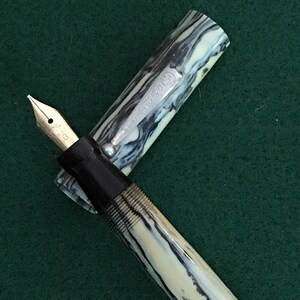 May include: A black and white marbled fountain pen with a silver band that says "AUTO-PENCIL" and a black cap. The pen is on a green surface.