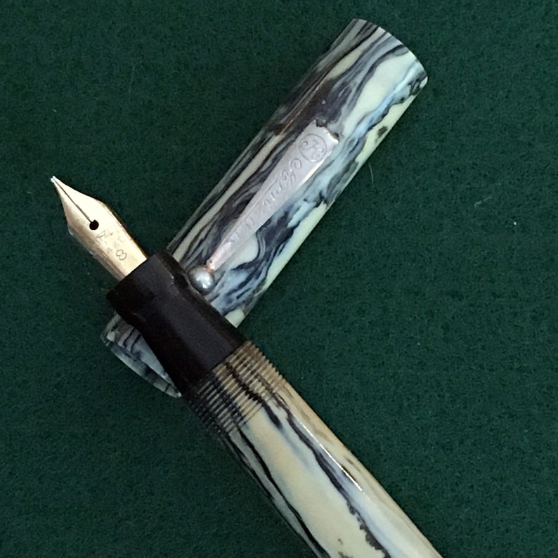 May include: A black and white marbled fountain pen with a silver band that says "AUTO-PENCIL" and a black cap. The pen is on a green surface.