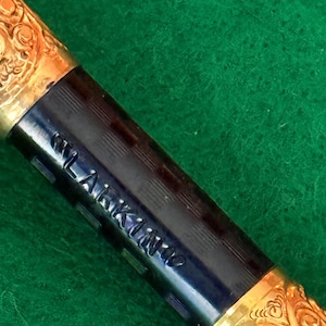 Puede incluir: Primer plano de una pluma estilogr&aacute;fica vintage. La pluma presenta un cuerpo azul oscuro con el nombre "LARKINS" grabado y detalles de filigrana dorada en el capuch&oacute;n y la secci&oacute;n de la pluma. La pluma descansa sobre una superficie verde.