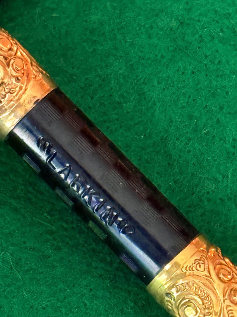 Puede incluir: Primer plano de una pluma estilogr&aacute;fica vintage. La pluma presenta un cuerpo azul oscuro con el nombre "LARKINS" grabado y detalles de filigrana dorada en el capuch&oacute;n y la secci&oacute;n de la pluma. La pluma descansa sobre una superficie verde.
