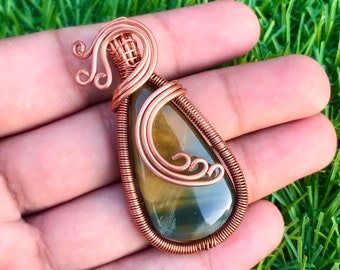 May include: A handcrafted pendant featuring a teardrop-shaped tiger's eye gemstone, wrapped in copper wire. The wirework includes decorative swirls and a bail for a necklace. The gemstone displays bands of gold, brown, and green.
