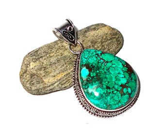 Può includere: Un ciondolo a forma di goccia con una vibrante pietra turchese, accentuata da motivi marroni. La pietra è incastonata in una montatura in metallo color argento con dettagli intricati. Il ciondolo è esposto su una piccola roccia grigia su sfondo bianco.