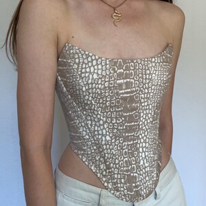 Può includere: Un top a corsetto senza spalline con un motivo a coccodrillo beige e bianco. Il top ha una scollatura curva e una silhouette aderente. La modella indossa pantaloni colorati chiari e una collana con pendente a serpente dorato.