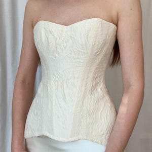 May include: Cream-colored strapless corset with textured fabric and a sweetheart neckline. A white shell necklace is worn around the neck. The corset is paired with a white skirt.