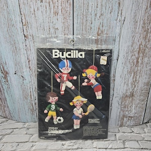 May include: A package of Bucilla Jeweled Holiday Ornaments, set of 4. The package features four felt ornaments depicting children playing sports. The text "IDEALLY SUITED TO CREATE A UNIQUE MOBILE" is also visible.