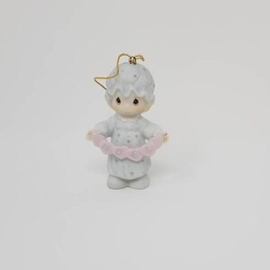 Puede incluir: Figura de cerámica de un niño con camisón, sosteniendo una pancarta rosa que dice "NOEL". La figura tiene un lazo dorado para colgar. La figura es azul claro y blanca.