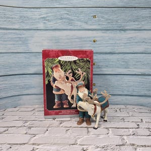 Könnte beinhalten: Ein Weihnachtsschmuck mit dem Weihnachtsmann und einem Rentier sowie eine Schachtel mit demselben Bild. Die Santa-Figur trägt einen blauen Mantel und Hut, braune Stiefel und hält die Zügel des Rentiers. Das Rentier hat einen blauen Sattel.