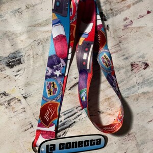 May include: A colorful lanyard with a repeating pattern of cassette tapes, bottles, and other graphics. The lanyard has a metal clasp and a tag that reads "la conecta" in black text on a blue gradient background.