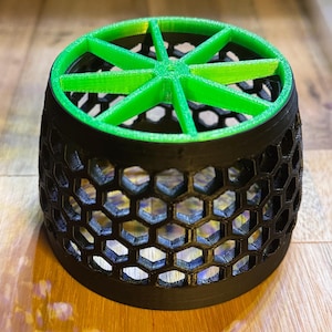 May include: A 3D-printed cylindrical object with a black honeycomb pattern on the sides and a green, star-shaped design on top. The object is sitting on a wooden surface.
