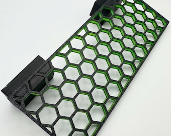 May include: A rectangular, honeycomb-patterned object with a black frame and green interior. The object has two black, rectangular extensions on the sides. The hexagonal pattern is evenly spaced throughout the design. The object is likely a decorative or functional item.