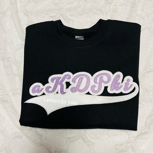 May include: Black sweatshirt with a white and purple "a KD Phi" applique in a baseball-style font. The applique has a white outline and the text "Letters by Jolie" is below it.