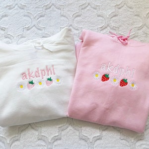 May include: Two hooded sweatshirts, one white and one pink, with the embroidered text "akdphi" and strawberry and daisy designs.