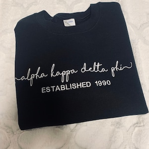 May include: Black sweatshirt with white embroidered text that reads "alpha kappa delta phi" and "ESTABLISHED 1990".
