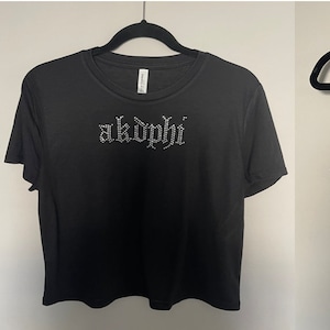 May include: Three black crop tops with the word "akdphi" in silver rhinestones on the front. The tops are on hangers.