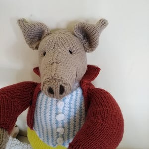 May include: Hand-knitted pig doll wearing a red jacket, a blue and white striped vest, and yellow pants. The pig has large ears and a long snout. The doll is made of soft yarn and has button details.