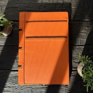 May include: A rectangular orange portfolio case featuring a smooth, solid-colored exterior with two horizontal black zippered pockets on the front and a vertical black zippered closure along the left side edge.