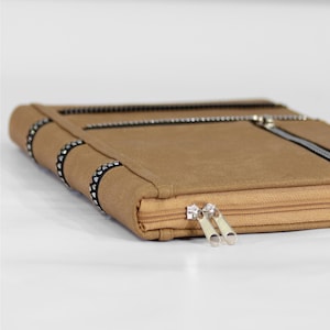 May include: A tan-colored, rectangular case with a zipper closure and silver-toned zipper pulls. The case features decorative black and silver accents along the edges and a zipper that runs across the top. The case appears to be made of a soft, felt-like material.