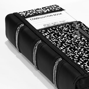 May include: Black zippered pouch with a composition book design. The pouch has a zippered pocket on the front with the text "COMPOSITION BOOK" printed on it. The pouch is made of a black fabric with a zippered closure.
