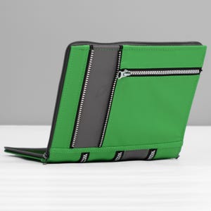 May include: Green and gray zippered case with a modern design. The case features a bright green exterior with a vertical gray stripe and multiple silver zippers. The case is open, revealing a glimpse of the interior.