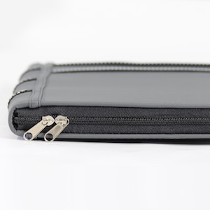 May include: A rectangular gray case featuring a black zipper closure with two silver-toned metal pull tabs. The case has a smooth, matte surface texture and a stitched seam running along the side edge, providing a clean and structured appearance.