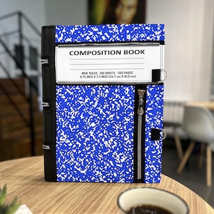 May include: A composition book cover with a blue and white speckled design. The cover has a black zipper and a black spine. The text "COMPOSITION BOOK" is printed on a white label. The dimensions are 9.75 inches x 7.5 inches.