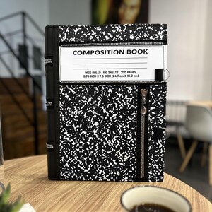 Może przedstawiać: Czarno-biały zeszyt kompozycyjny z zapięciem na zamek błyskawiczny. Okładka ma nakrapiany wzór, widoczny jest napis "COMPOSITION BOOK". Wymiary: 24,7 cm x 19,0 cm.
