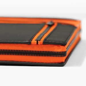 Puede incluir: Primer plano de una bolsa de cuero negro con cremallera y ribete naranja. La cremallera es visible y la bolsa está doblada por la mitad.
