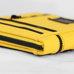 May include: A bright yellow rectangular pouch featuring multiple black zippered compartments. The exterior is constructed with smooth yellow material and accented by thick black plastic zippers that run horizontally across the front and along the side edge.