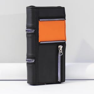 May include: A black wallet with an orange panel and silver zippers. The wallet is designed to resemble a book, with vertical lines and a zipper closure. The wallet is made of leather-like material.