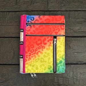 May include: A rectangular notebook cover featuring a vibrant rainbow gradient with a leopard print pattern. It includes a solid pink spine, two horizontal zippered compartments, and one vertical zippered pocket, all with silver-toned metal zippers.