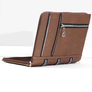 May include: A brown leather portfolio with multiple silver zippers. The portfolio is open, revealing a dark brown interior. The zippers are black and silver, adding a contrasting detail to the overall design. The portfolio is designed to hold documents.