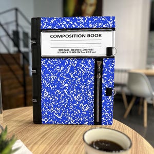 May include: A blue and white speckled composition book cover featuring a black spine with zipper details. The front displays a white label with the text COMPOSITION BOOK, WIDE RULED, 100 SHEETS, 200 PAGES, 9.75 INCH X 7.5 INCH (24.7 cm X 19.0 cm).