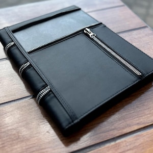 May include: A black rectangular portfolio or notebook cover featuring a smooth surface with three prominent silver-toned metal zips. One zip runs vertically along the front pocket, while two additional zips are positioned horizontally.