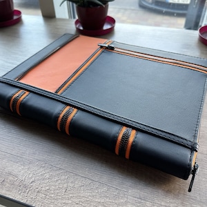 Könnte beinhalten: Ein schwarzes Leder-Zipper-Portfolio mit orangefarbenen Reißverschluss-Akzenten. Das Portfolio ist geschlossen und liegt auf einer Holzoberfläche.