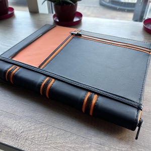 May include: A black leather zippered notebook with orange zipper accents. The notebook is closed and lying on a wooden surface.