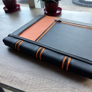 May include: A rectangular portfolio case featuring a combination of black and orange panels. The exterior is accented with black zippers that run vertically along the spine and horizontally across the top section, creating a structured design.