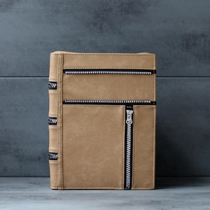 May include: A tan rectangular notebook cover featuring a smooth matte finish. The front panel is accented with two horizontal black zippers and one vertical black zipper with a silver pull. The spine displays three additional black zipper details.