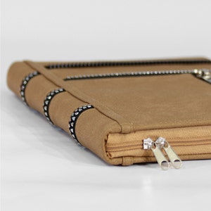 May include: A tan-colored rectangular case with a smooth, matte finish. The case features a side zipper closure with two silver-toned metal pulls and decorative black bands accented with small, repeating silver-toned heart-shaped studs.
